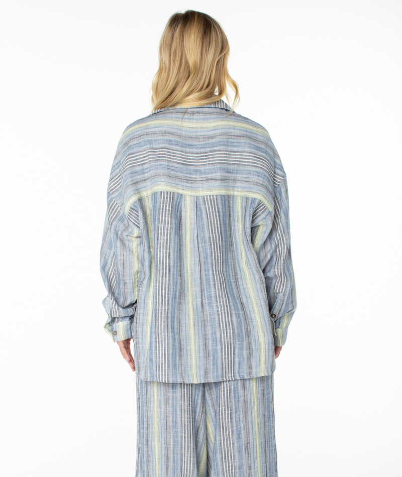 A person with long blonde hair, facing away, wears the Roxy Lets Get Away Top—a loose, long-sleeved shirt in blue, yellow, and gray vertical stripes on a white background. Its oversized fit creates an effortless look that could easily double as a stylish beach cover up.