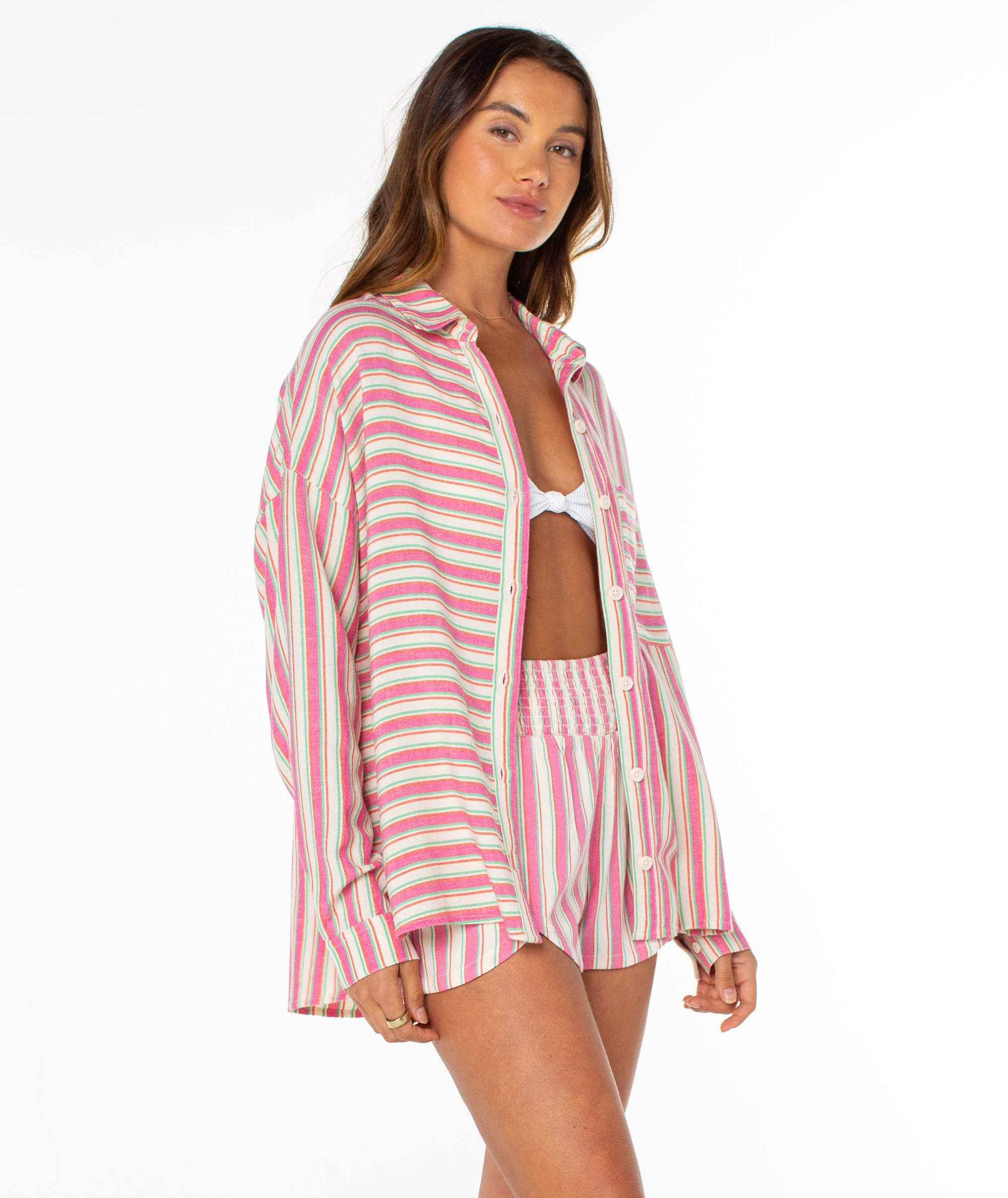 A woman models the Roxy Lets Get Away Top—a loose, long-sleeve, pink and white striped shirt—styled over matching shorts and a bikini top, standing against a plain white background.