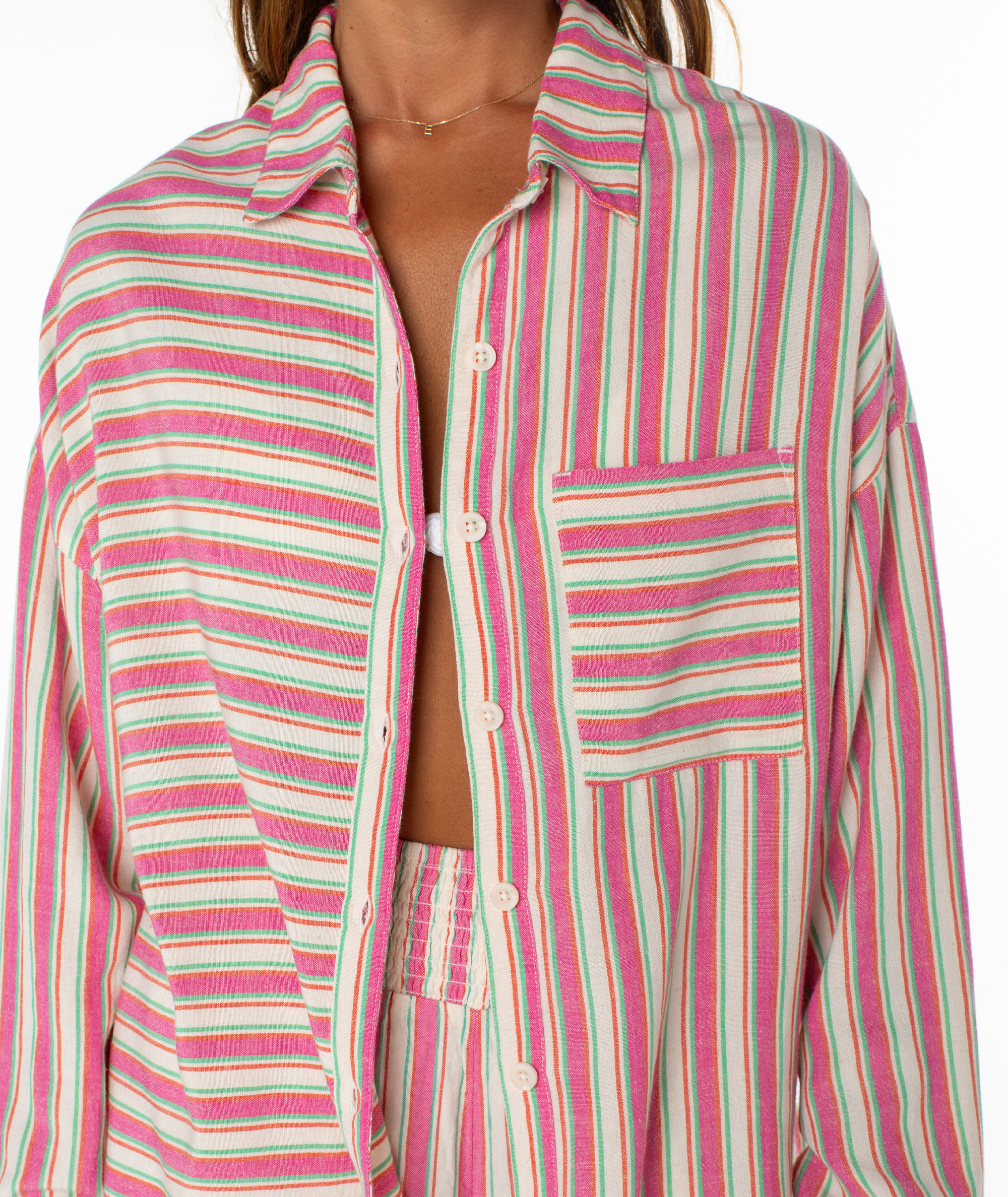 Wearing the Lets Get Away Top by Roxy—a vibrant, vertically striped shirt in pink, green, and cream with a stylish front pocket—someone leaves it slightly open to reveal a matching top underneath.