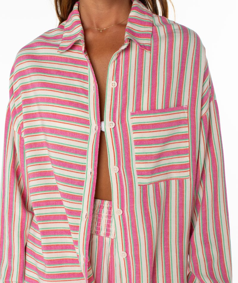 Wearing the Lets Get Away Top by Roxy—a vibrant, vertically striped shirt in pink, green, and cream with a stylish front pocket—someone leaves it slightly open to reveal a matching top underneath.