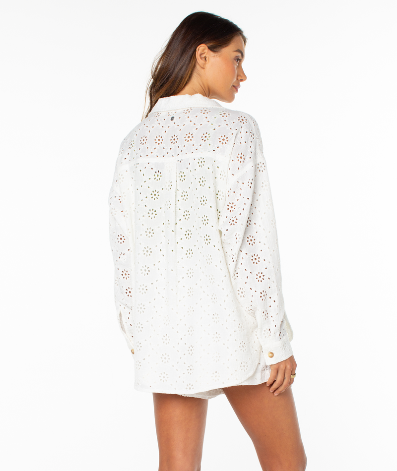 A woman faces away wearing the Roxy Morning Time Top—a loose white summer shirt with long sleeves, buttoned cuffs, and a floral cutout design. Its relaxed fit is ideal for a beachy feel. The background is plain white.