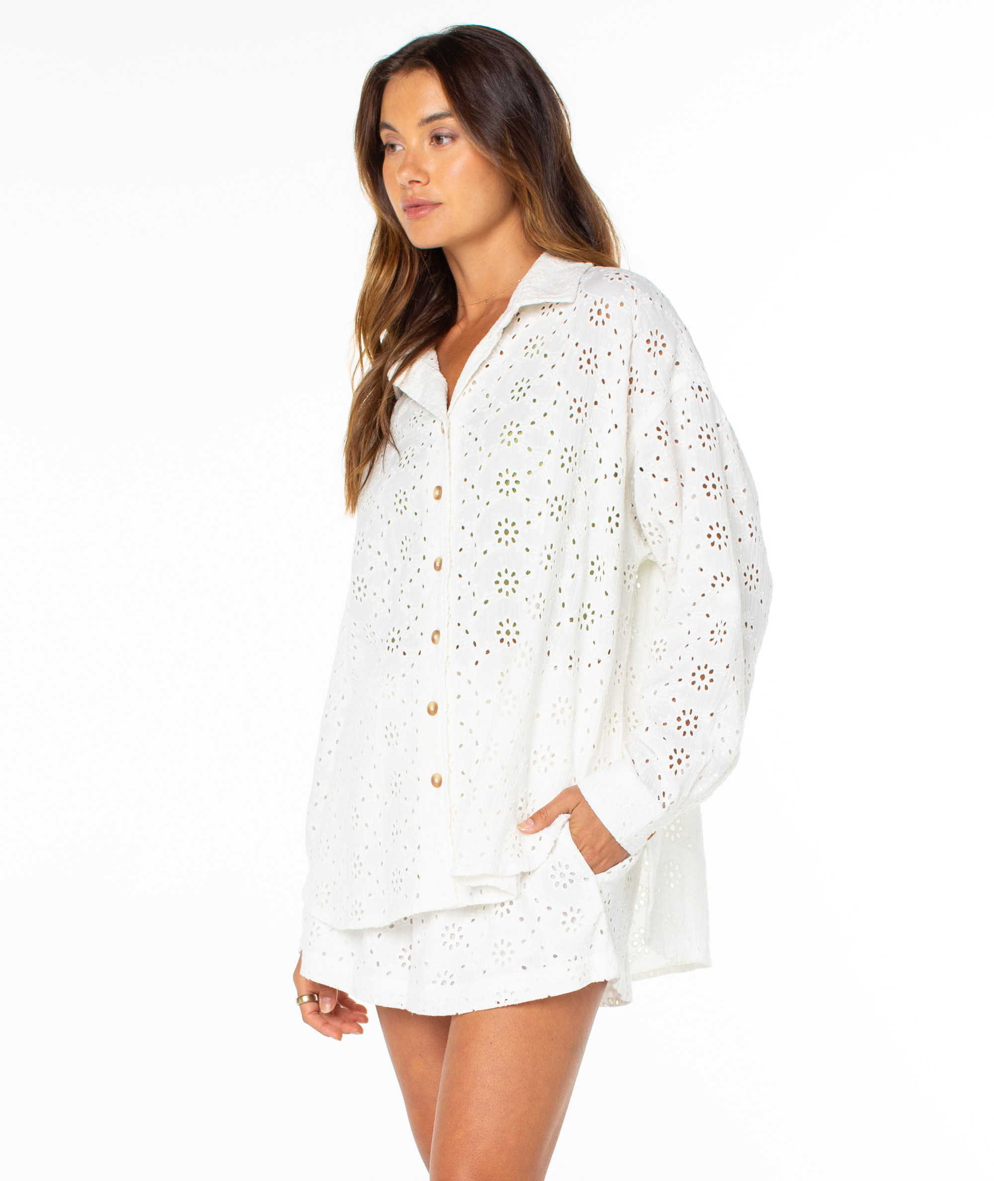 A woman wears the Roxy Morning Time Top, a loose-fitting white summer shirt, standing against a plain white background with one hand in her pocket and looking to the side.
