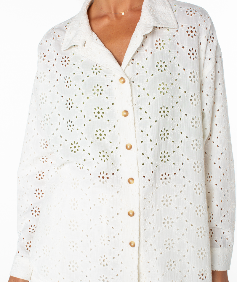 A person wears the Roxy Morning Time Top—a white, long-sleeve shirt with a floral eyelet pattern, gold buttons, and a collar. Made of lightweight, semi-sheer fabric, it’s perfect for warm summer days.