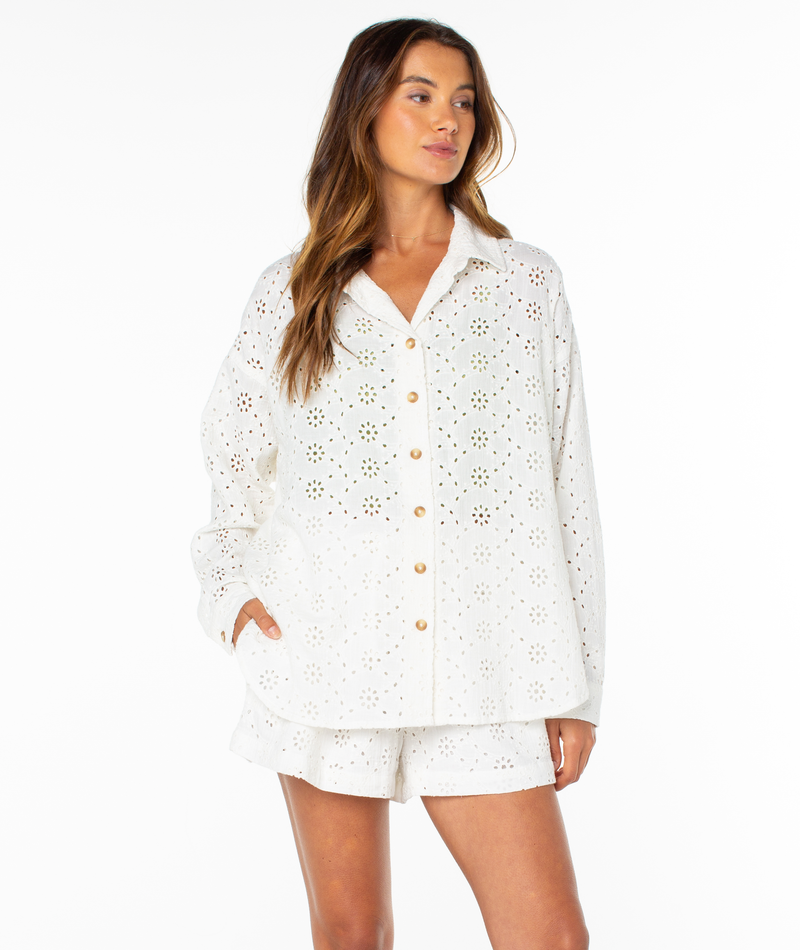 A woman models the Roxy Morning Time Top—a white, long-sleeve summer shirt—paired with matching eyelet shorts, standing with one hand in her pocket against a plain white background.