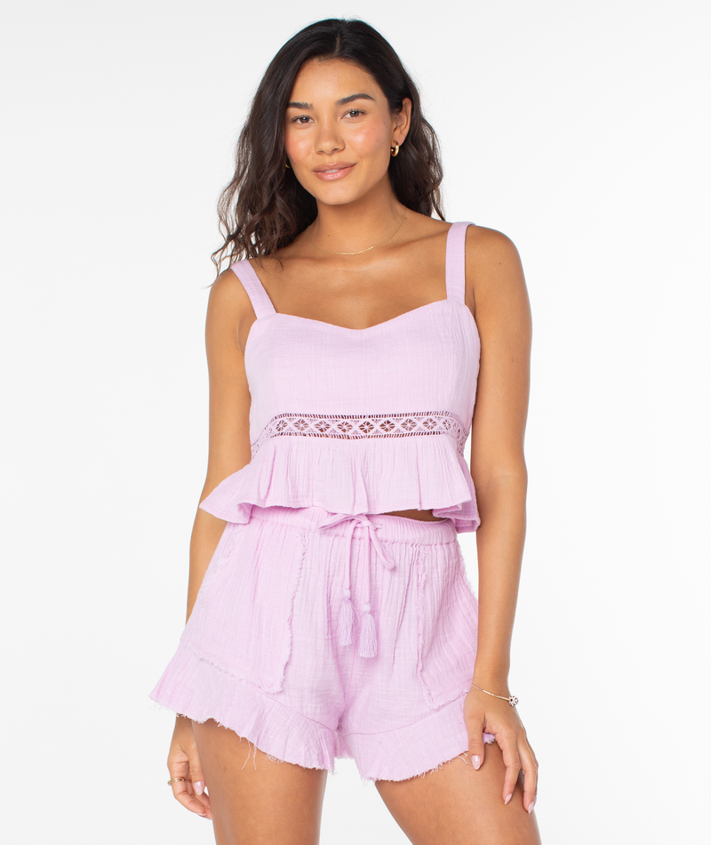 A woman wearing the Roxy Island Time Tank in Orchid Bouquet Solid, featuring a light pink camisole with a lace midriff detail and matching shorts with a drawstring waist.