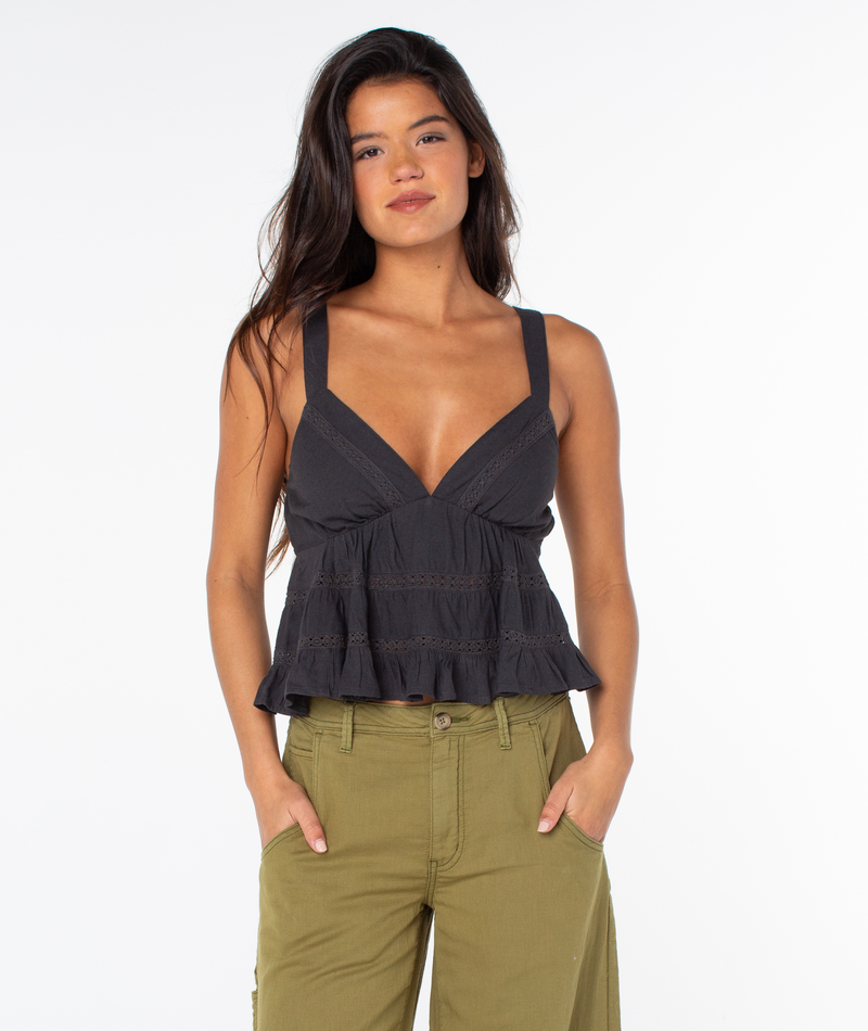 A woman with long dark hair wears a sleeveless black Roxy Layla Tank featuring a deep V neckline, paired with olive green linen blend pants, standing with her hands in her pockets against a plain white background.