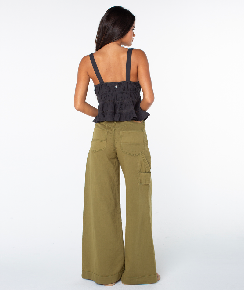 A woman stands barefoot, facing away, wearing the Roxy Layla Tank—a black, sleeveless top featuring a V neckline and ruffled accents—paired with wide-leg olive green linen-blend pants with pockets. The background is plain white.