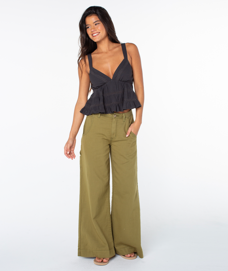 A woman stands smiling against a plain white background, wearing the Roxy Layla Tank—a sleeveless black top featuring a flattering V-neckline and ruffled waist—paired with olive-green wide-leg pants. She has one hand in her pocket.