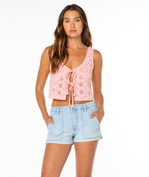A woman with long brown hair wears the Roxy Kayla Tank, a pink crochet eyelet top with front ties and a scoop neckline, paired with light blue denim shorts. She stands with hands in her pockets against a plain white background.