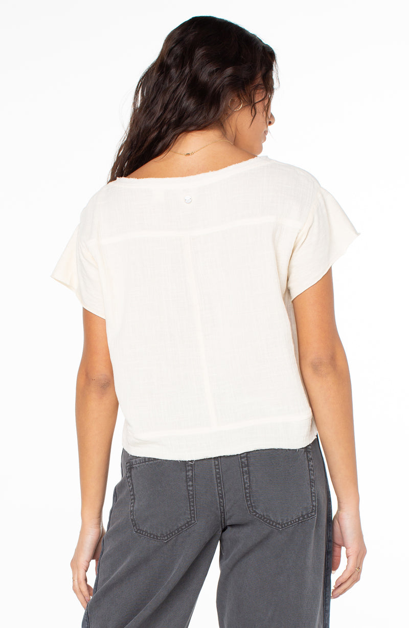 Back view of the cream-colored Malina Top, a short-sleeved shirt with a raw-edged neckline and a single button detail at the center back.