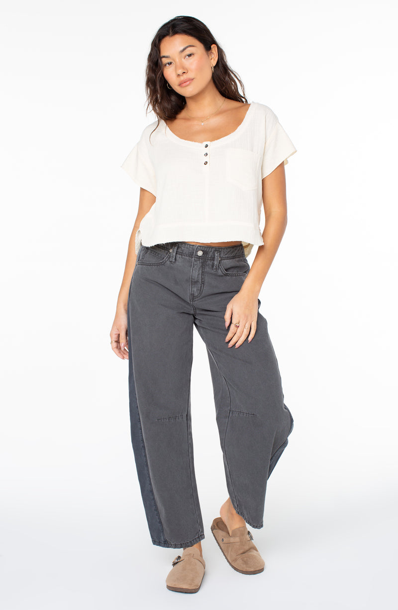 A woman wears a cream-colored, short-sleeved Roxy Malina Top with a button-front neckline, paired with grey wide-leg jeans and tan clogs.