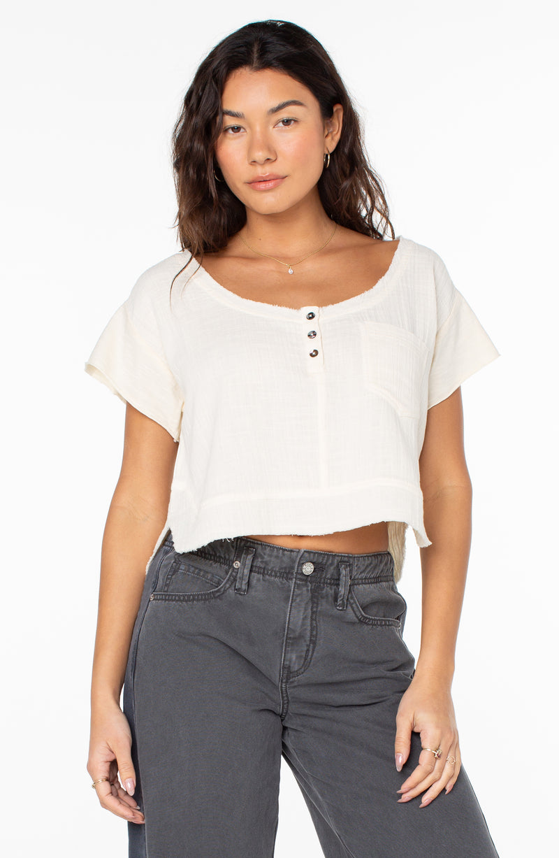 A woman wears the Roxy Malina Top, a parchment solid, cream-colored, cropped short-sleeve shirt featuring a front button placket, a chest pocket, and a raw-edge hem.