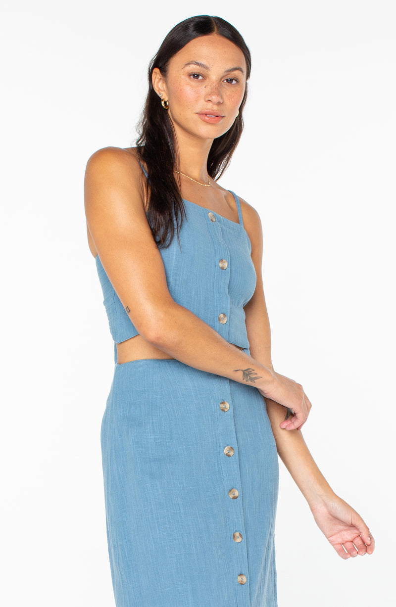 A person wears a blue button-down Island Nomad Top with thin straps and a matching button-down midi skirt.