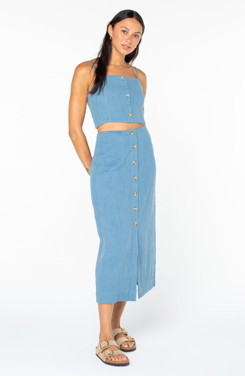 A woman wearing a blue button-front Roxy Island Nomad Top with a matching midi-length skirt and sandals.