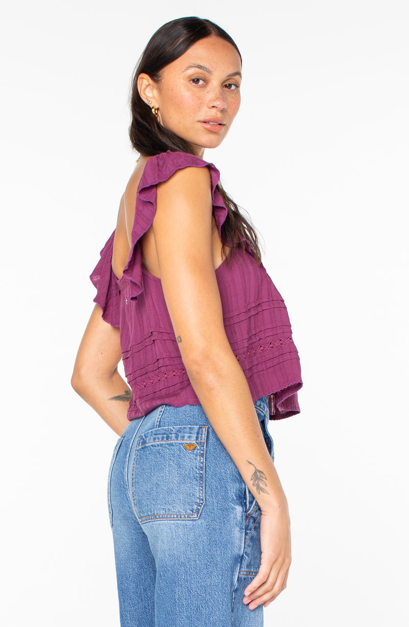 A person wearing a purple, sleeveless, ruffled-shoulder Roxy Coastal Rays Top and blue jeans.