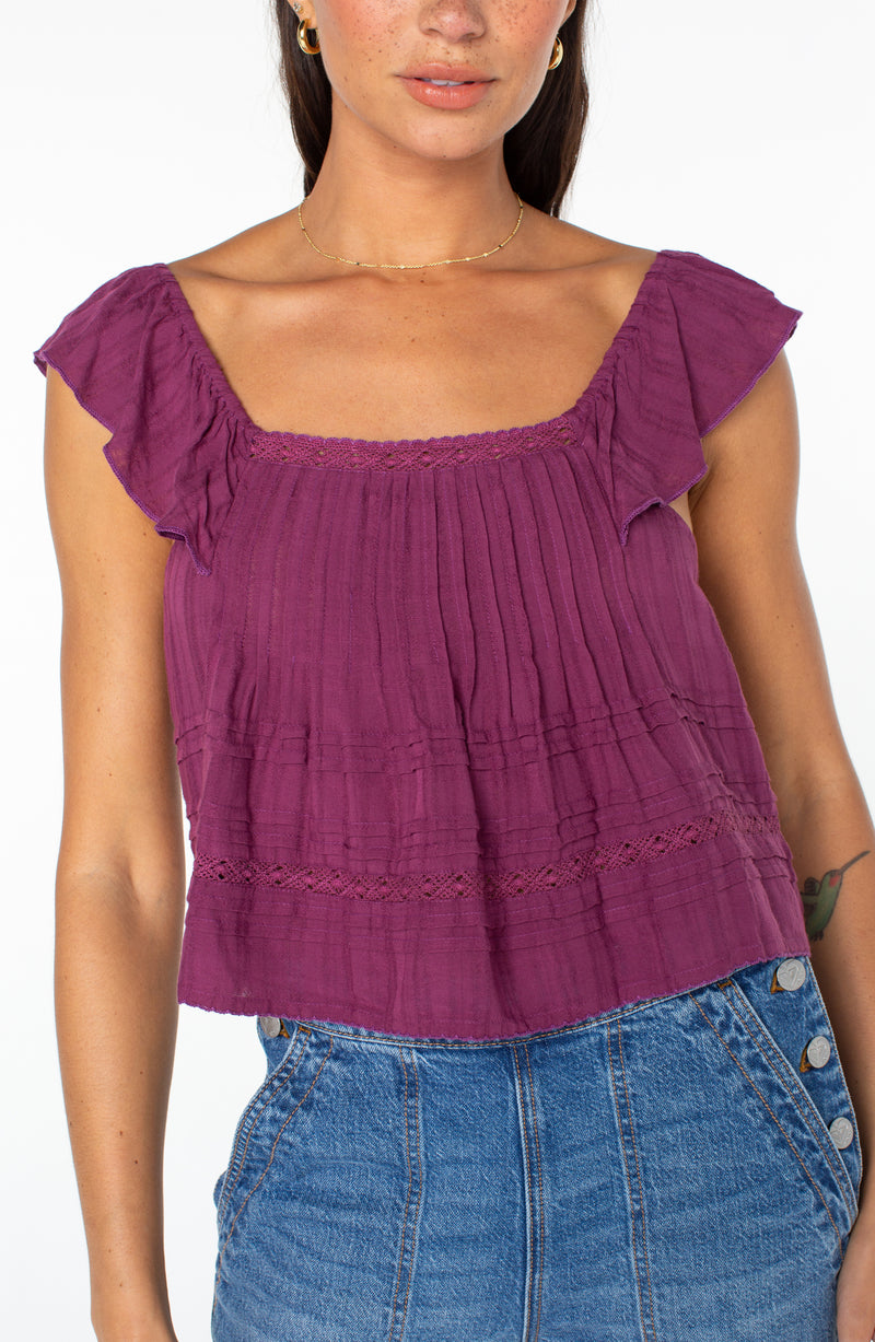 A plum-colored Roxy Coastal Rays Top with ruffled cap sleeves, a square neckline, and horizontal lace detailing, worn with blue jeans.