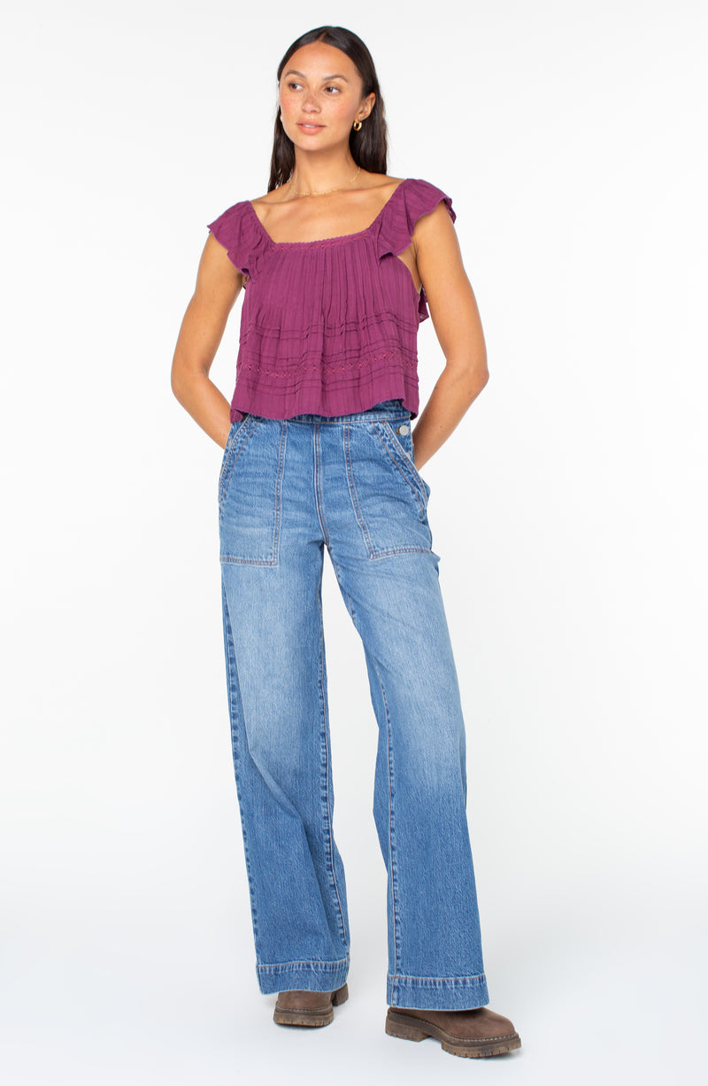 A person wearing a plum-colored ruffled sleeveless Coastal Rays Top and wide-leg blue jeans.