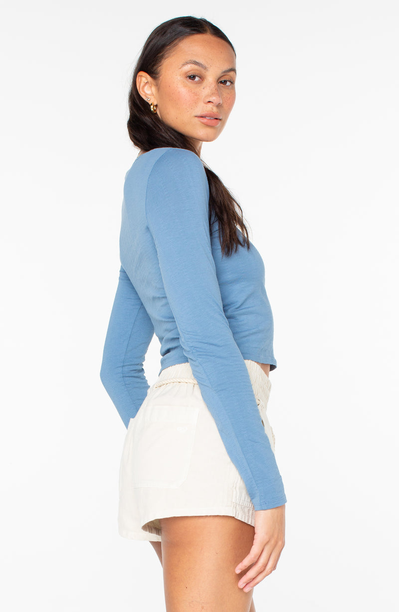 A person wearing a blue Roxy Cuddle Up Long Sleeve crop top and light-colored shorts.