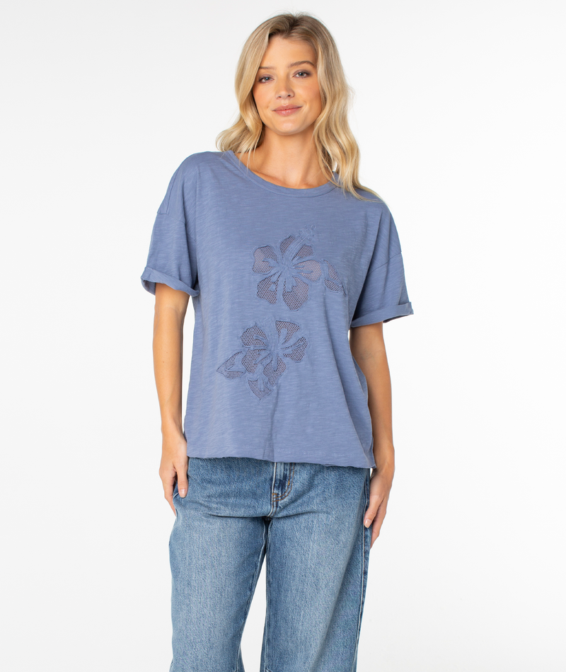 A person wearing the Roxy Sunset Sessions Tee in Wild Wind Hibiscus Cutout, paired with blue jeans, stands against a plain white background, smiling at the camera.