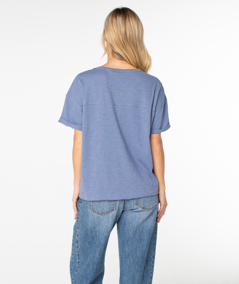 A person with long blonde hair, wearing the Roxy Sunset Sessions Tee in a loose boxy fit and blue jeans, stands facing away from the camera against a plain white background.