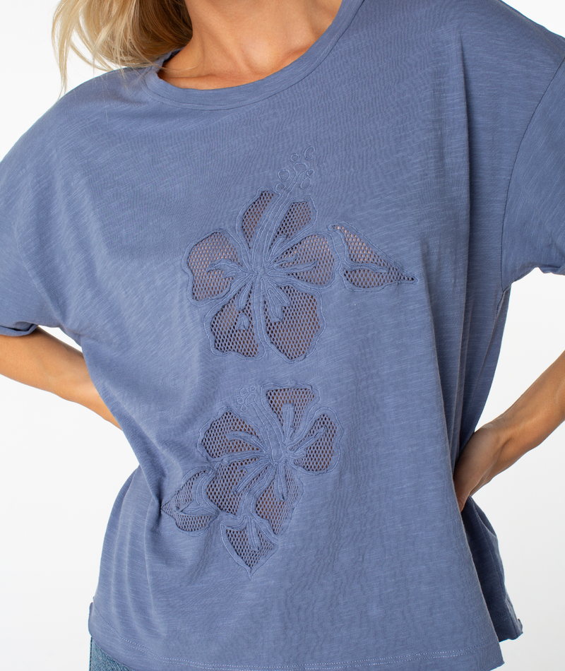 A person wearing the Roxy Sunset Sessions Tee—a loose, short-sleeved blue top featuring mesh floral cutout applique designs on the front. Only the upper torso is visible, with one hand resting on the hip.
