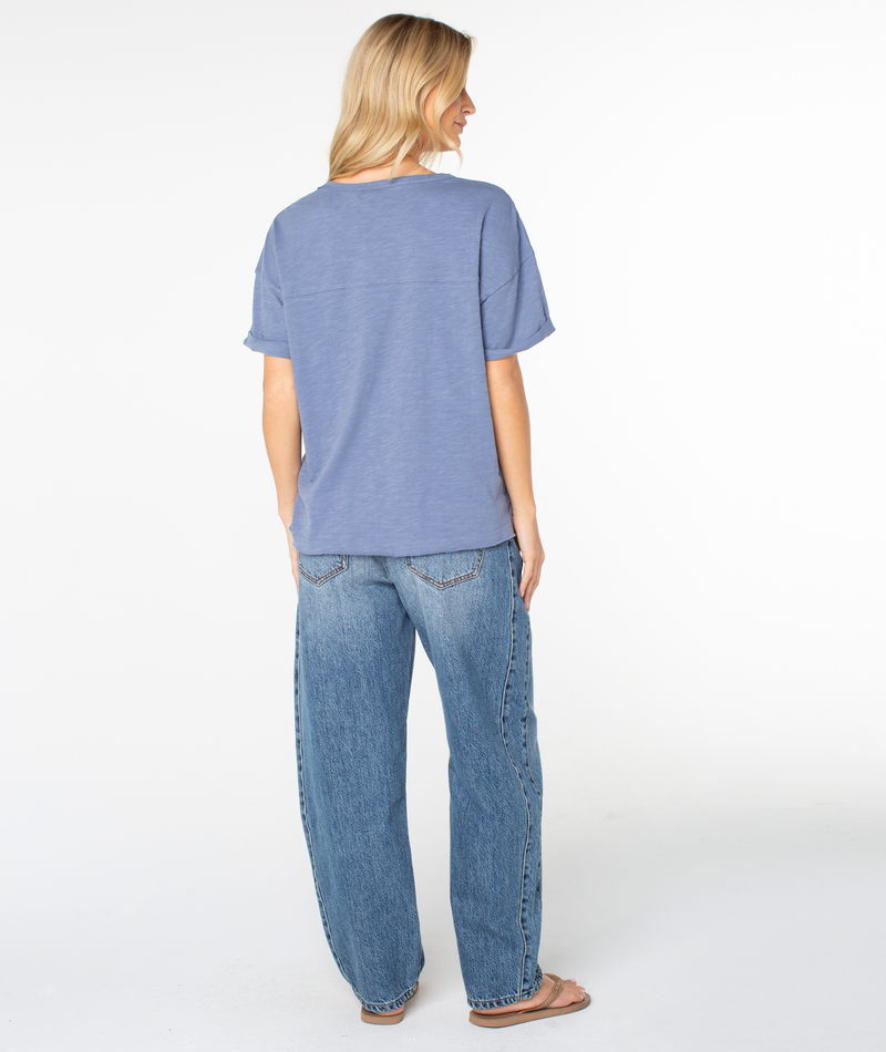 A person stands facing away, wearing the Roxy Sunset Sessions Tee in blue, paired with loose blue jeans and sandals, against a plain white background.