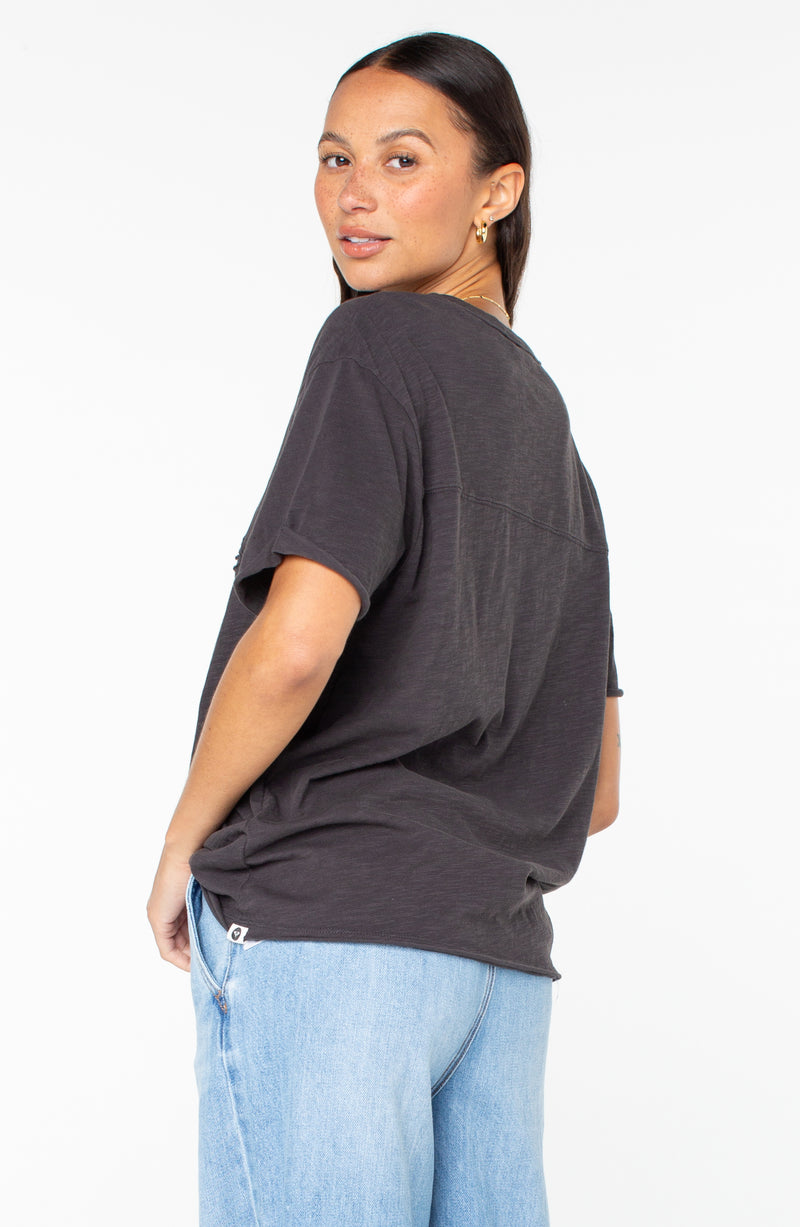 A person wearing a charcoal Sunset Sessions Tee and light-wash jeans, seen from behind while looking over their shoulder.