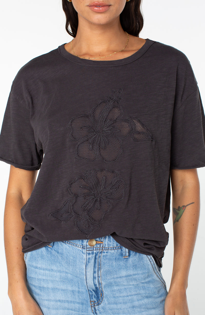 Roxy Sunset Sessions Tee in dark gray with short sleeves and two embroidered hibiscus flower cutouts on the front, tucked into light-wash denim jeans.