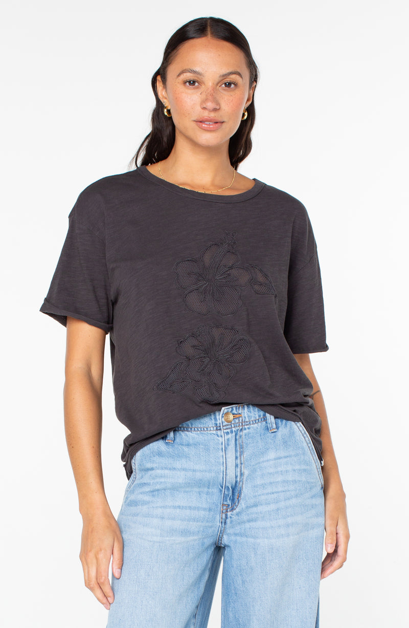 A charcoal gray Roxy Sunset Sessions Tee with two large hibiscus-shaped cut-outs on the front, worn with light-wash blue jeans.
