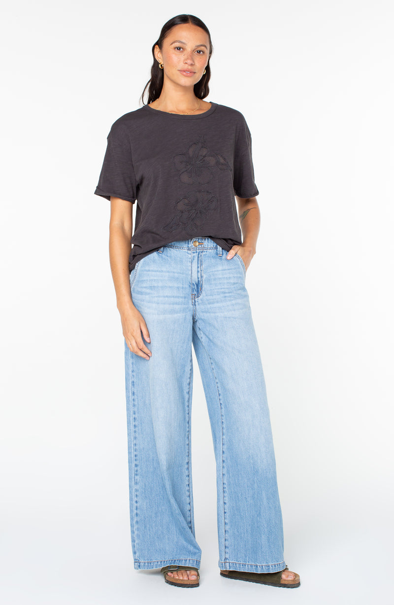 A person wears a charcoal grey Sunset Sessions Tee featuring subtle tonal floral embroidery paired with light-wash wide-leg denim jeans.