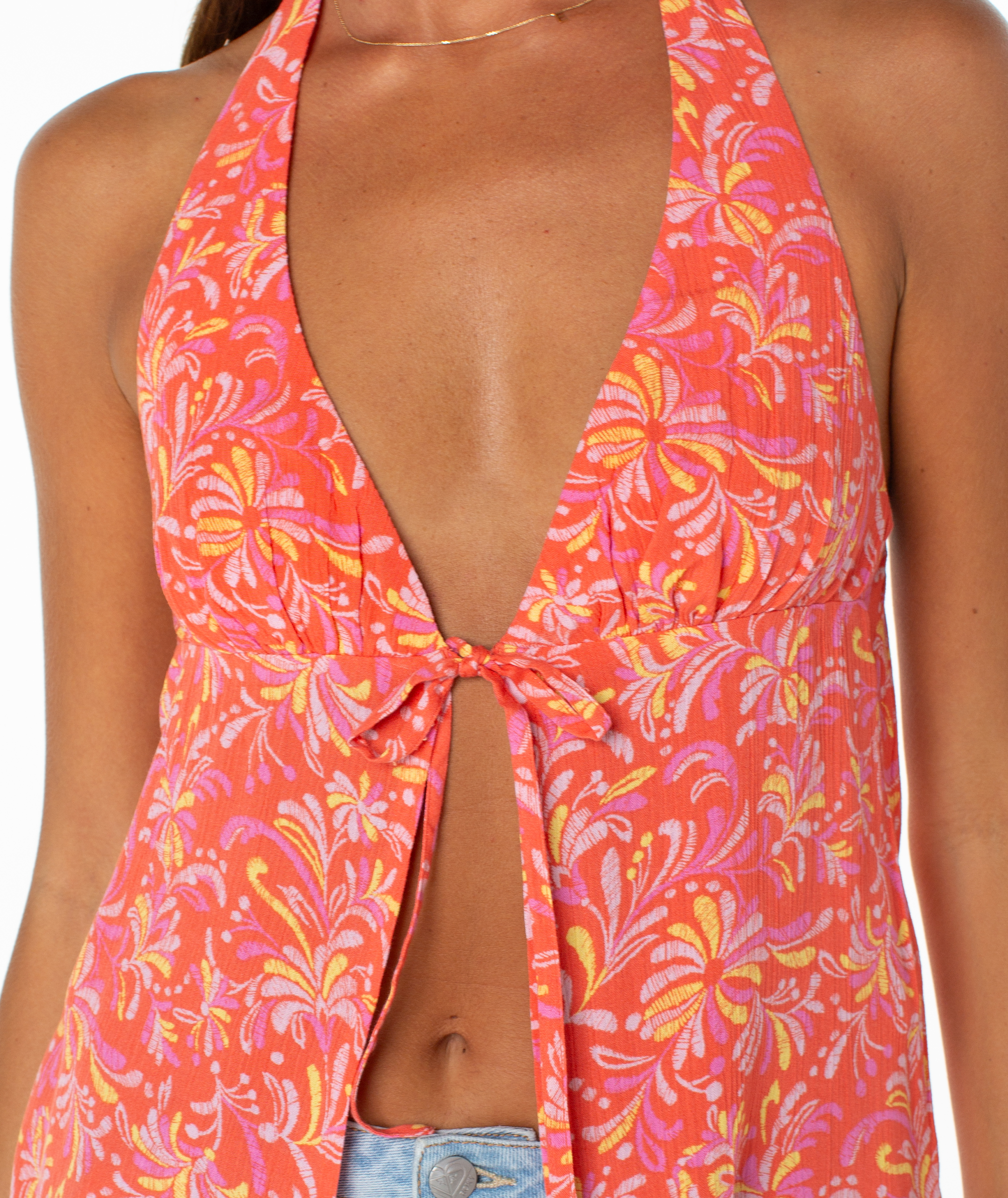 A woman wears the Roxy Fairweather Top—a sleeveless halter with a deep V neckline and pink, orange, and yellow floral pattern tied at the bust—paired with blue jeans. Only her upper torso is visible.