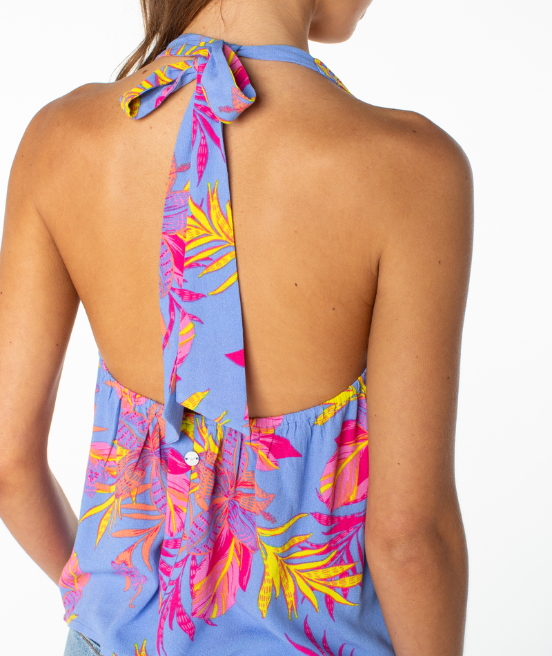 A person wears the Roxy Fairweather Top—a blue halter with a pink and yellow tropical floral print, tied at the neck in retro beachwear style, featuring an open back and baring part of the arm and shoulder.