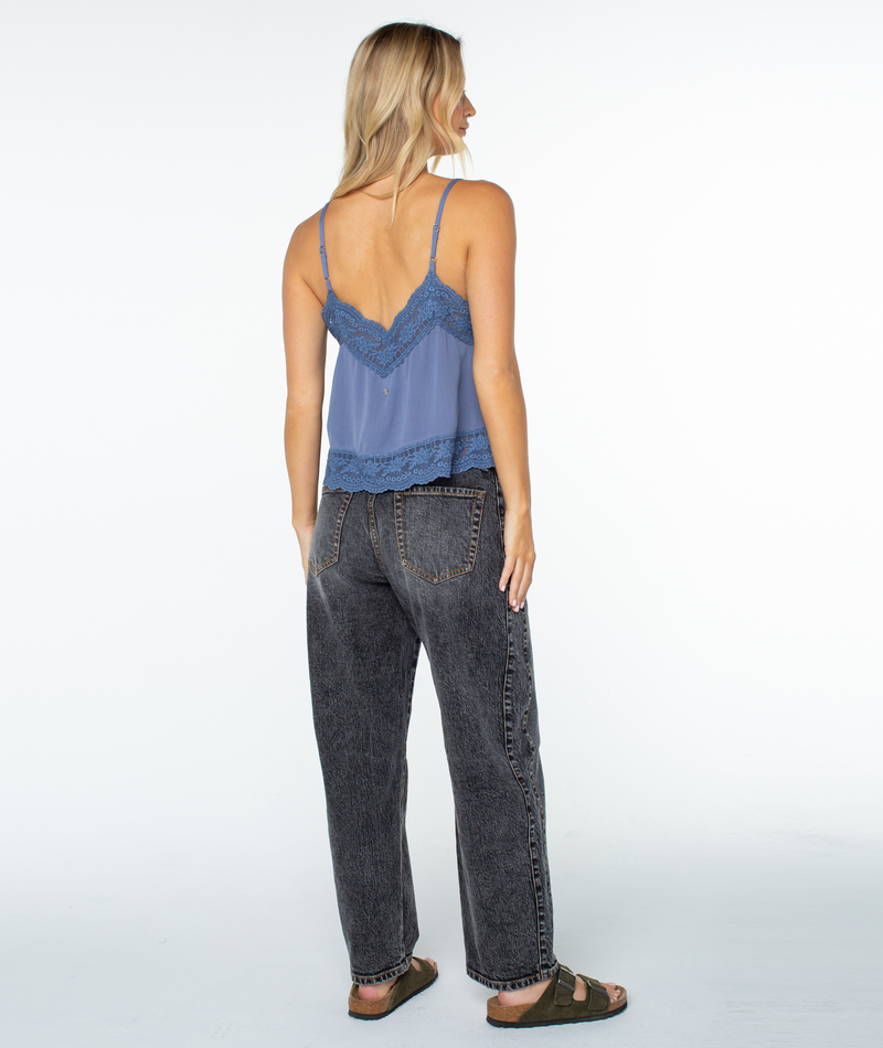A woman faces away, wearing the Roxy Shadow Side Top—a blue lace-trimmed camisole with a scalloped V-neck—paired with black washed jeans and sandals, her straight blonde hair set against a white background.