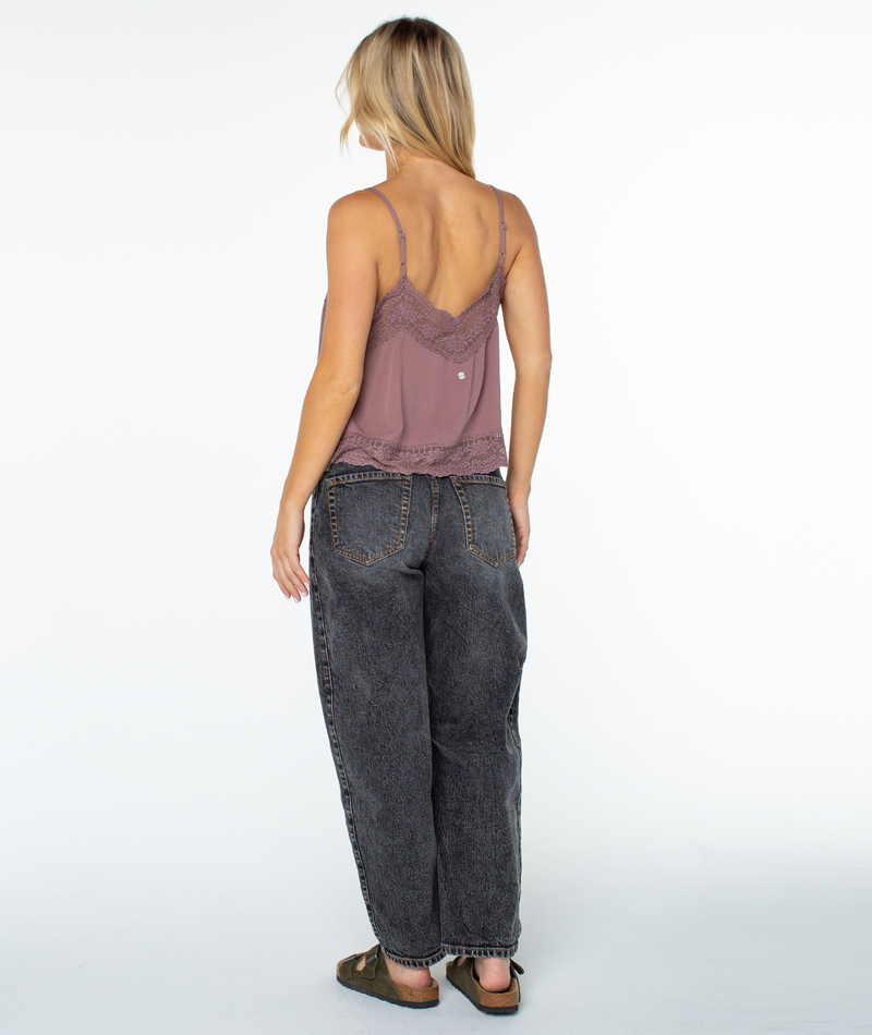 A woman with blonde hair, wearing the Roxy Shadow Side Top, loose black jeans, and sandals, stands facing away against a plain white background.
