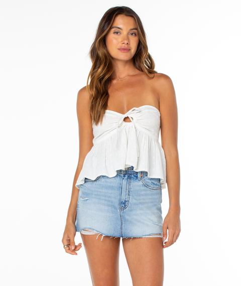 A woman wears the Roxy Ventura Tube Top—a white strapless style with a front knot and keyhole—paired with a light blue frayed denim mini skirt, standing against a plain white background.