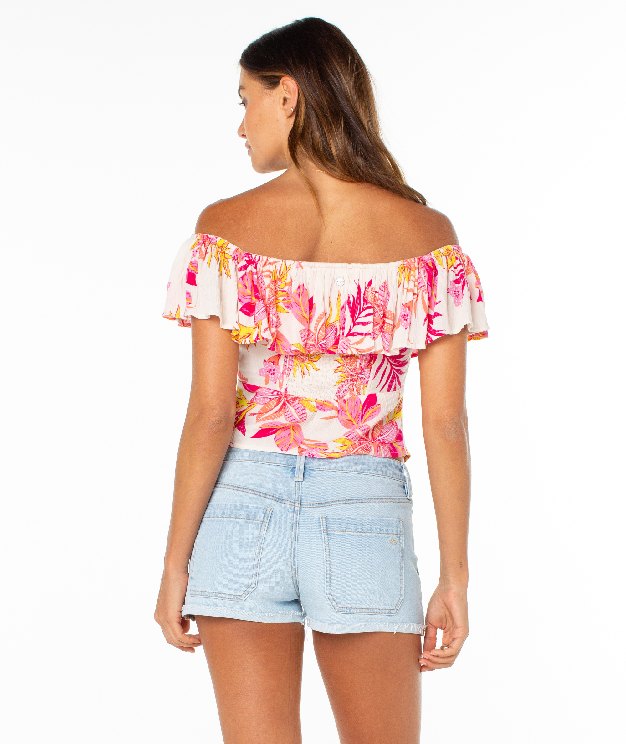 A woman with long brown hair stands facing away against a white background, wearing the Roxy Clear Sky Top—an off-the-shoulder floral blouse with pink, orange, and yellow patterns—and light blue denim shorts.