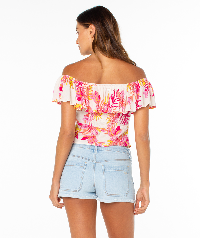 A woman with long brown hair stands facing away against a white background, wearing the Roxy Clear Sky Top—an off-the-shoulder floral blouse with pink, orange, and yellow patterns—and light blue denim shorts.