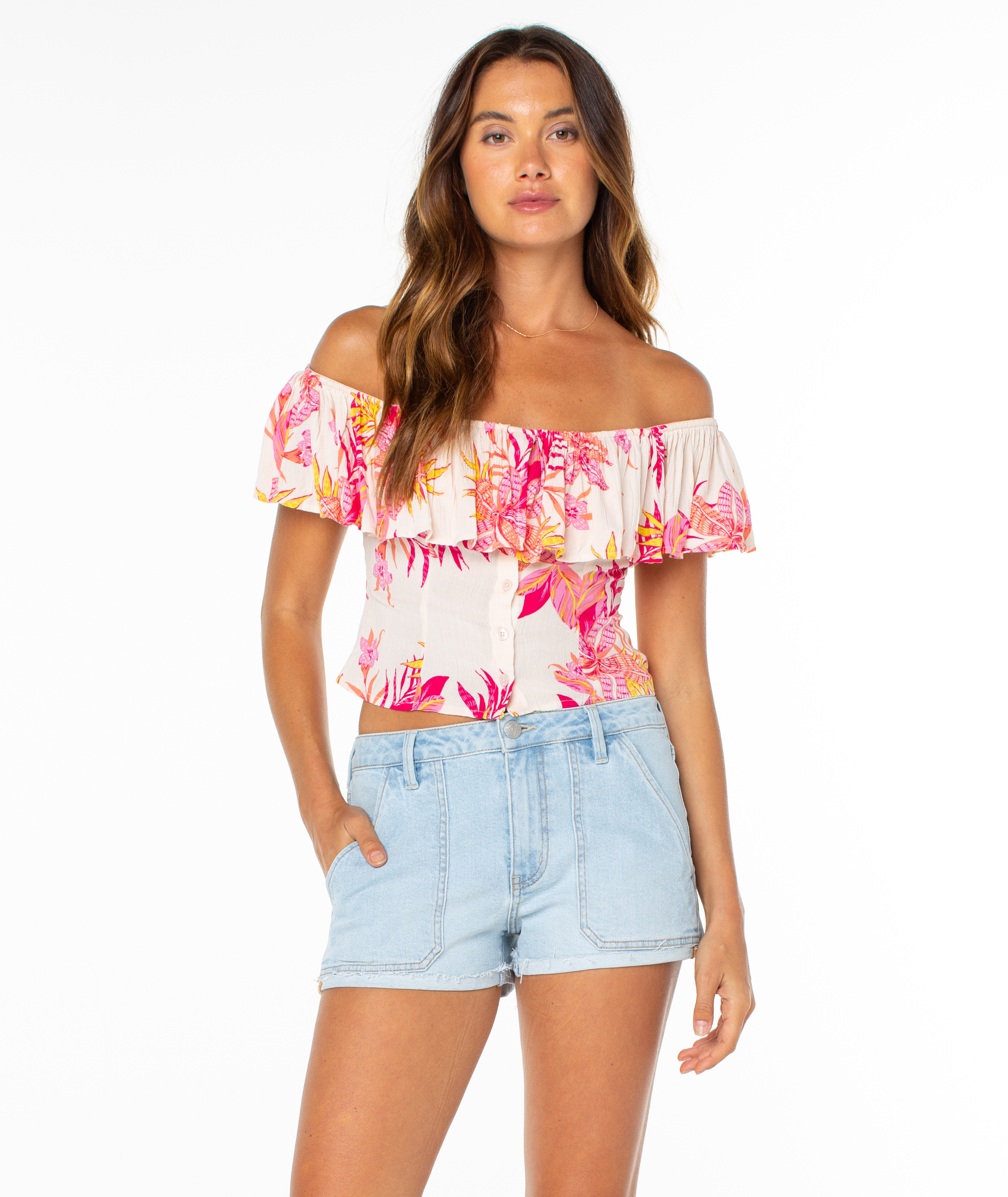 A woman stands against a white background, wearing the Roxy Clear Sky Top—an off-the-shoulder pink and orange floral blouse—paired with light blue denim shorts. She has long, wavy brown hair and a relaxed, confident look.