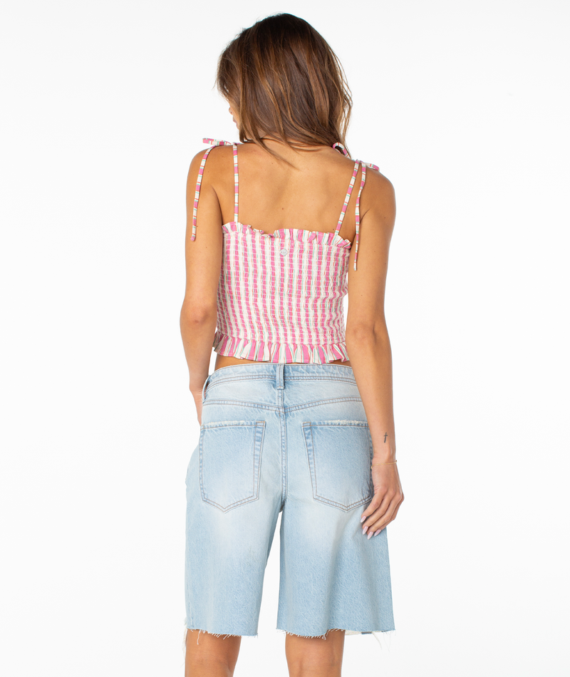 A woman is seen from the back in the Roxy Avalon Tube Top, a smocked pink and white striped sleeveless style with shoulder ties, paired with light-wash knee-length denim shorts. The background is plain white. Perfect for summer casual looks.