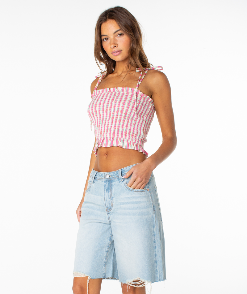 A woman with long brown hair poses against a white background wearing the Roxy Avalon Tube Top in pink and white, paired with light blue frayed-hem denim shorts. She has a relaxed, summer-ready look.