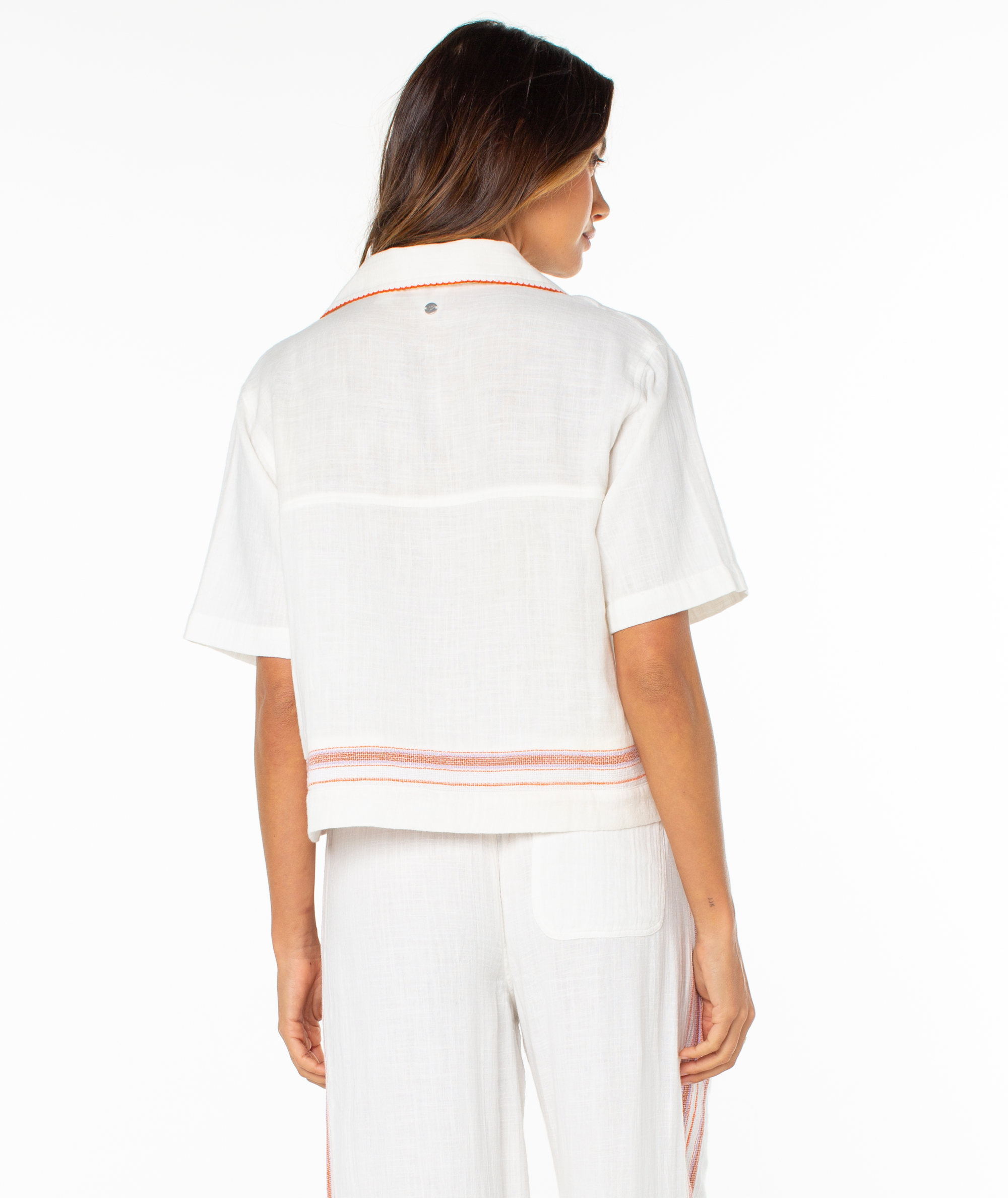 A woman faces away, wearing Roxy’s Fiesta Siesta Shirt—a summery striped top with red accents at the hem—paired with white pants against a plain white background.