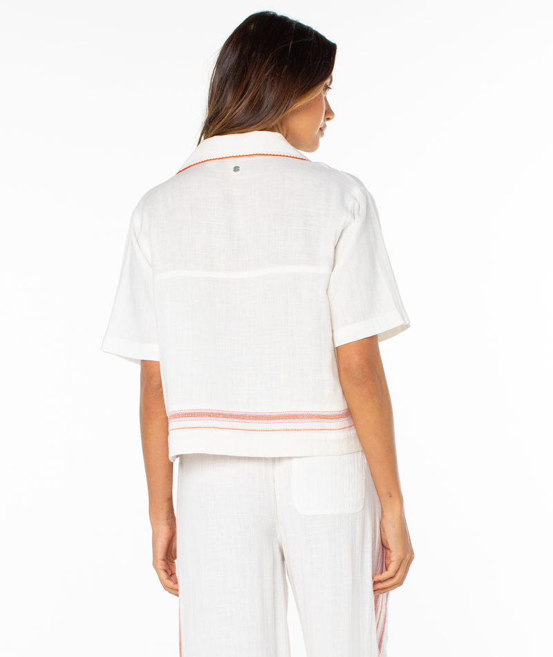 A woman faces away, wearing Roxy’s Fiesta Siesta Shirt—a summery striped top with red accents at the hem—paired with white pants against a plain white background.
