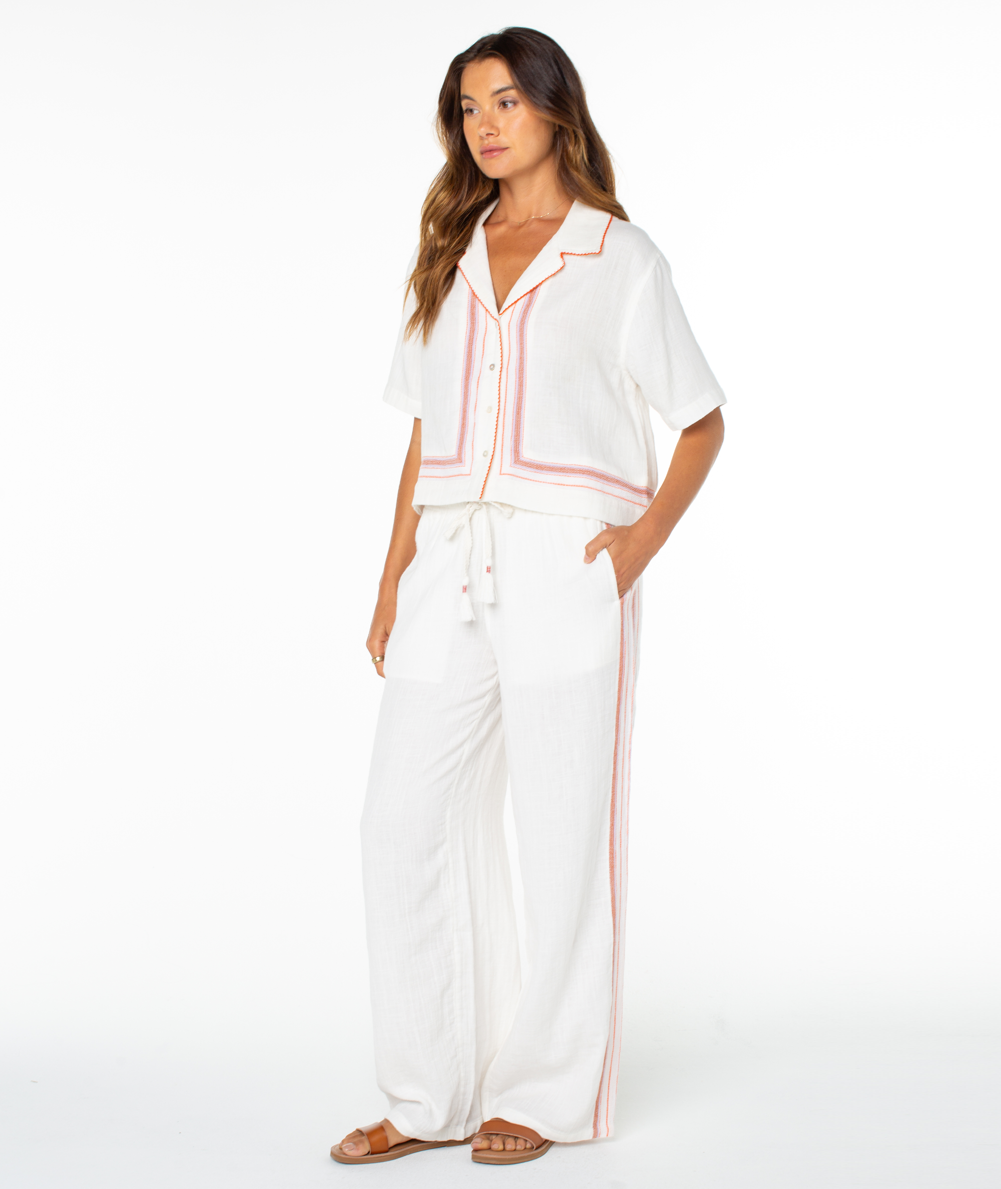 A woman models the Roxy Fiesta Siesta Shirt—white, loose-fitting with red-orange trim—and matching wide-leg pants and brown sandals, shown against a plain white background.