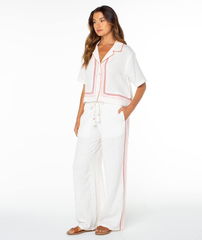 A woman models the Roxy Fiesta Siesta Shirt—white, loose-fitting with red-orange trim—and matching wide-leg pants and brown sandals, shown against a plain white background.