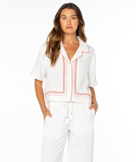 A woman with long brown hair models the Roxy Fiesta Siesta Shirt, a white top with orange trim, and matching drawstring pants. She stands hands in pockets against a white background, highlighting the set's easygoing style.
