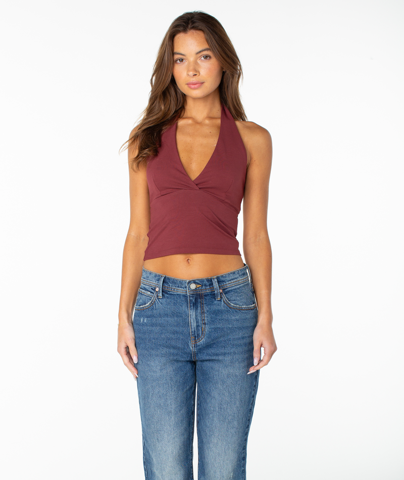 A woman with long brown hair wears the Roxy South Shore Halter Tank in Port Solid—a sleeveless, cropped-fit top—paired with blue jeans, standing against a plain white background and looking at the camera with a neutral expression.