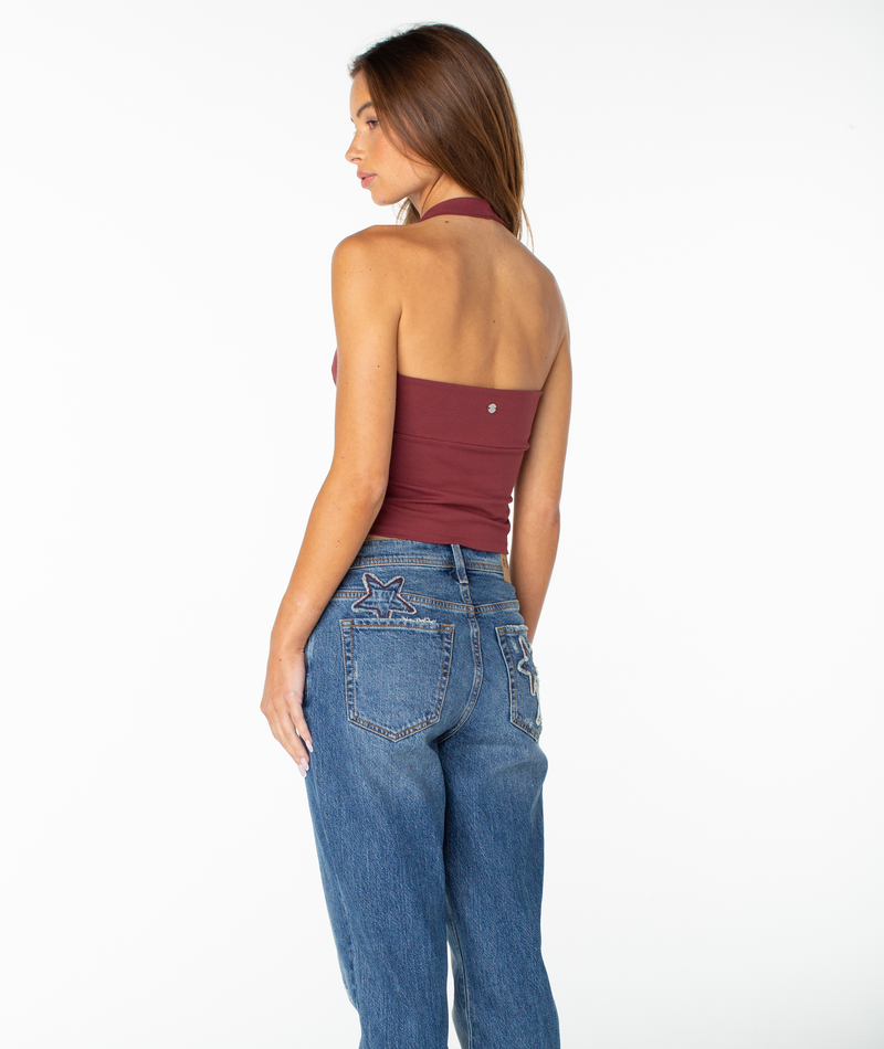A woman stands with her back to the camera, wearing the Roxy South Shore Halter Tank in a maroon cropped fit and blue jeans featuring star patches on the back pockets, channeling a subtle retro style against a plain white background.