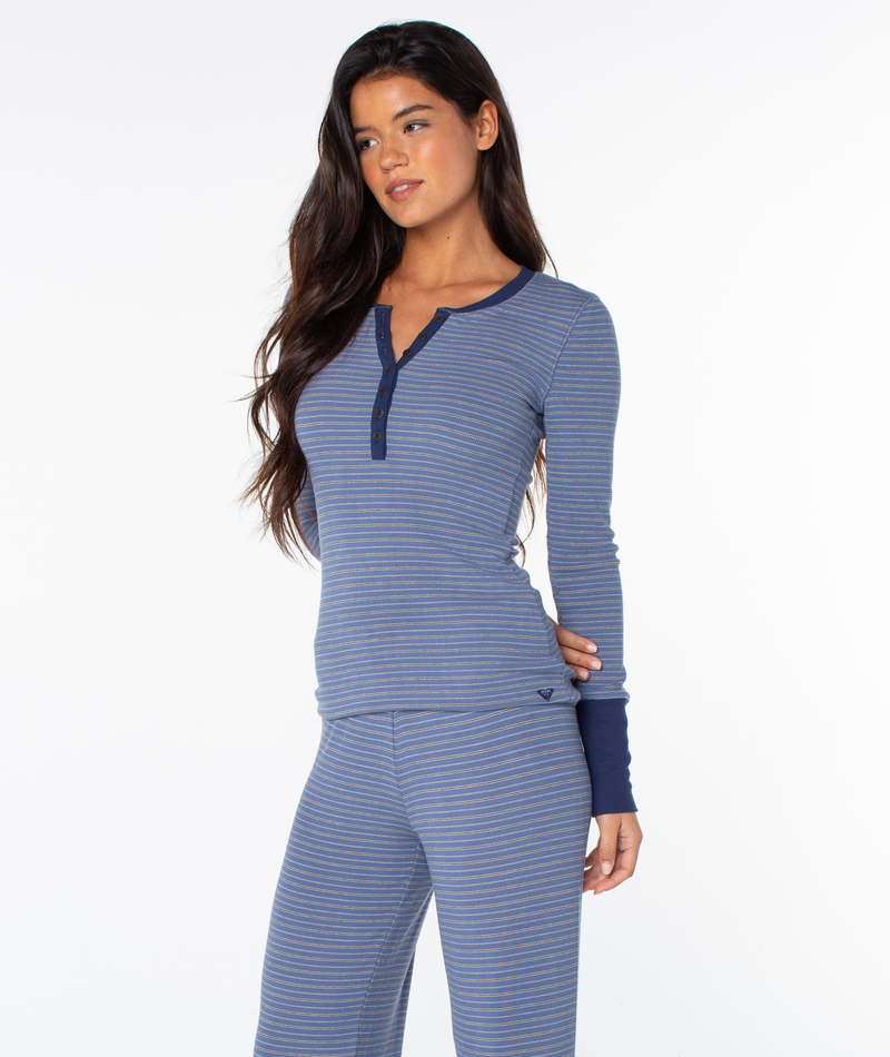 A woman with long dark hair models the Roxy Hillside Long Sleeve Henley Top in blue and gray stripes and matching pants, standing against a plain white background.