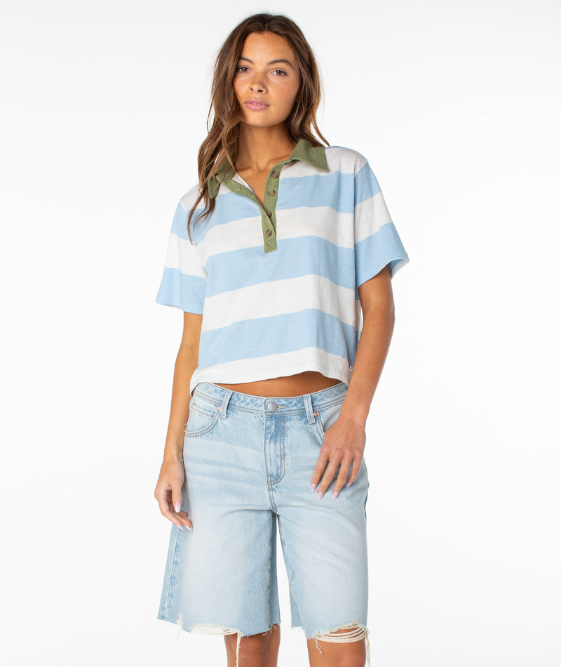 A woman wearing the Brookview Polo by Roxy in Cerulean Rugby Stripe, paired with light denim knee-length shorts, stands against a white background.