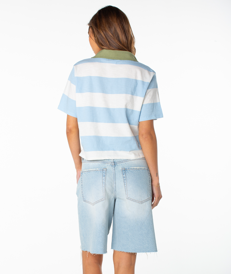 A person with long hair stands facing away, wearing the Roxy Brookview Polo in a relaxed fit with light blue and white bold stripes, paired with light blue denim bermuda shorts. The background is plain white.